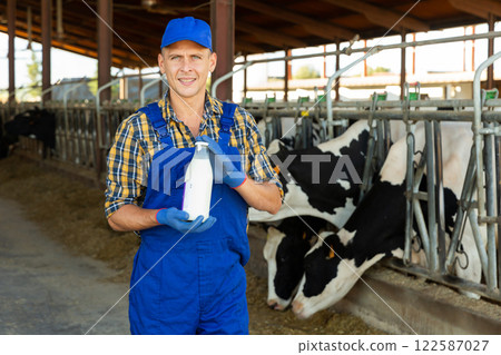 Portrait of dairy farm worker with bottle of milk in his hands against background of cows in stall 122587027