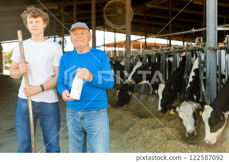 Positive aged man dairy farm owner standing with teen grandson near stalls with cows Positive aged man dairy farm owner standing with teen grandson near stalls with cows 122587092