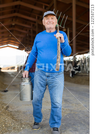 Elderly male breeder with milk can and pitchfork standing in stall on background with herd of cows on livestock farm Elderly male breeder with milk can and pitchfork standing in stall on background with herd of cows on livestock farm 122587113