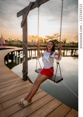 Asian woman poses cutely, sitting on a swing, on a wooden bridge, backlit photo. 122588525