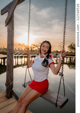 Asian woman poses cutely, sitting on a swing, on a wooden bridge, backlit photo. 122588526