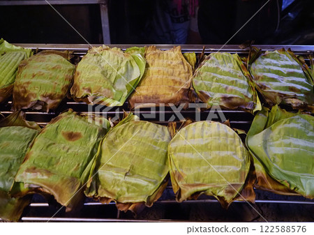 A Grilled banana leaf wrapped snacks on barbecue grill, showcasing traditional street food preparation. leaves are charred, indicating smoky flavor 122588576
