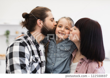 Caucasian mom and dad lovingly kiss young daughter in bright kitchen setting, showcasing joy and togetherness. 122588697