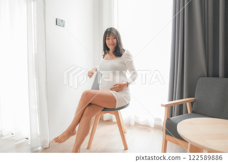 Pregnant Asian woman in white dress in bedroom Pregnant Asian woman in white dress in bedroom 122589886
