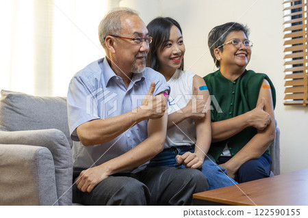 Senior Asian couple and teen woman get vaccinated with bandage on arm show thumb up sign in living room. Grandfather and grandmother get vaccine. 122590155