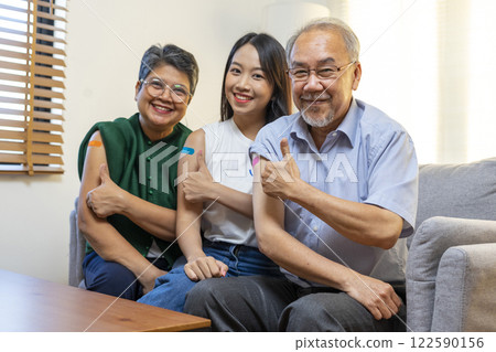 Senior Asian couple and teen woman get vaccinated with bandage on arm show thumb up sign in living room. Grandfather and grandmother get vaccine. 122590156