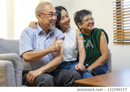 Senior Asian couple and teen woman get vaccinated with bandage on arm show thumb up sign in living room. Grandfather and grandmother get vaccine. 122590157