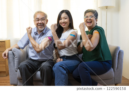 Senior Asian couple and teen woman get vaccinated with bandage on arm show thumb up sign in living room. Grandfather and grandmother get vaccine. Senior Asian couple and teen woman get vaccinated with bandage on arm show thumb up sign in living room. Grandfather and grandmother get vaccine. 122590158