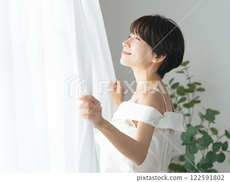 一位中年婦女拉開窗簾,露出清爽的笑容 一位中年婦女拉開窗簾,露出清爽的笑容 122591802