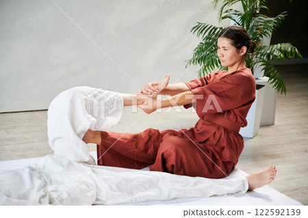Masseuse making thai yoga massage. Woman therapist doing traditional massage treatment, stretching female patient's body. Client lying on mat, enjoying hand massage in calm atmosphere. 122592139