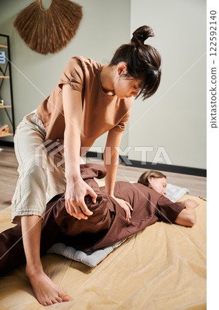 Masseuse making thai yoga massage. Woman therapist doing traditional massage treatment, stretching female patient's body. Client lying on mat, enjoying hand massage in calm atmosphere. 122592140