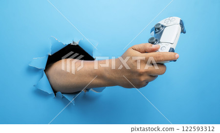 Human hand holding the gamepad, joystick, or video game controller through ripped paper. mockup or copy space. Concept of playing video games, entertainment or leisure activity 122593312