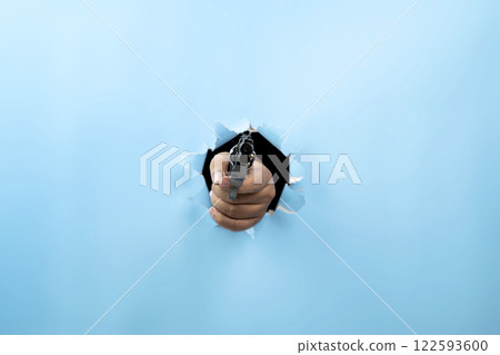 Human hand holding handgun through ripped paper. Hand gestures. Mockup or Copy space. Aiming weapons, war, crime, or violence 122593600