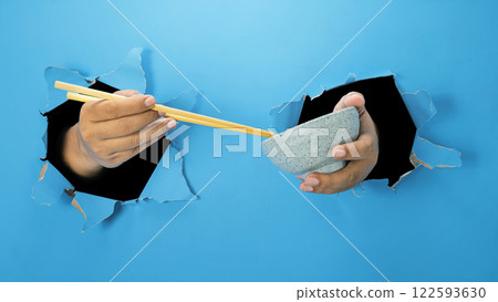 A human hand is holding a wooden chopstick and bowl through ripped paper. Hand gestures to reach or grab food. Mockup or Copy space 122593630