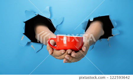A human hand is holding a cup through ripped paper. Hand gestures. Mockup or Copy space 122593642