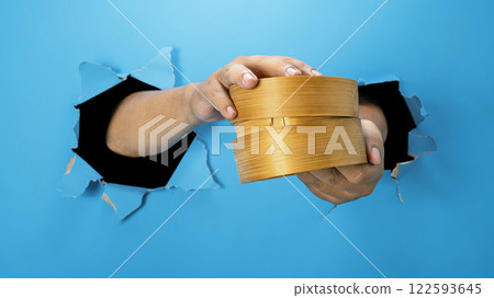 A human hand is holding a bamboo steamer through ripped paper. Hand gestures to serve food. Mockup or Copy space 122593645
