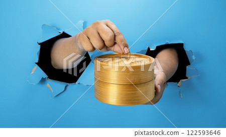 A human hand is holding a bamboo steamer through ripped paper. Hand gestures to serve food. Mockup or Copy space A human hand is holding a bamboo steamer through ripped paper. Hand gestures to serve food. Mockup or Copy space 122593646
