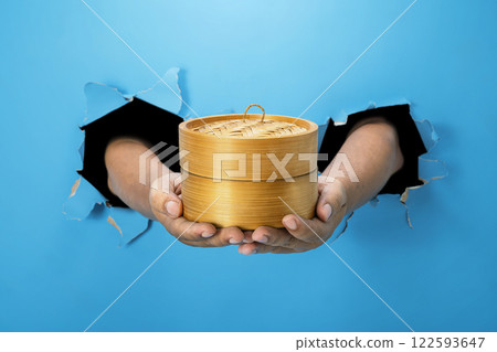 A human hand is holding a bamboo steamer through ripped paper. Hand gestures to serve food. Mockup or Copy space A human hand is holding a bamboo steamer through ripped paper. Hand gestures to serve food. Mockup or Copy space 122593647