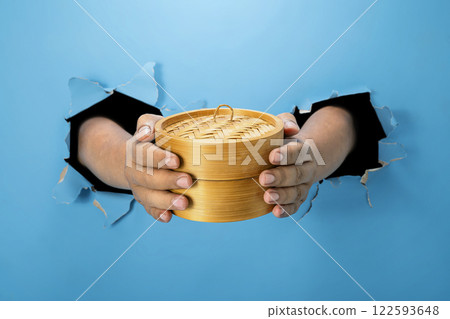 A human hand is holding a bamboo steamer through ripped paper. Hand gestures to serve food. Mockup or Copy space A human hand is holding a bamboo steamer through ripped paper. Hand gestures to serve food. Mockup or Copy space 122593648