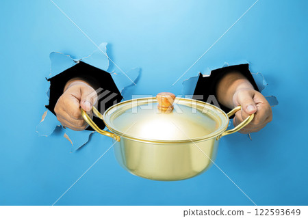 A human hand is holding a saucepan through ripped paper. Hand gestures to serve food. Mockup or Copy space A human hand is holding a saucepan through ripped paper. Hand gestures to serve food. Mockup or Copy space 122593649