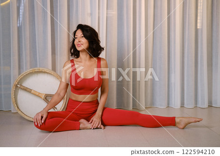 A woman in a red tracksuit sits with a drum in a gymnasium. Kalimeditation class 122594210
