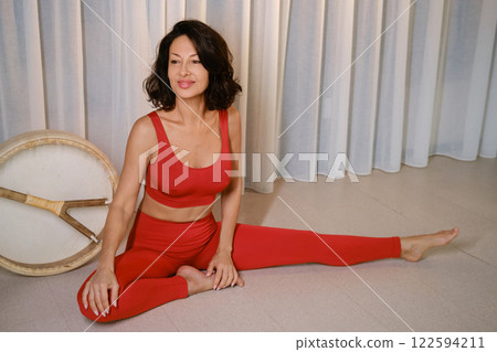 A woman in a red tracksuit sits with a drum in a gymnasium. Kalimeditation class A woman in a red tracksuit sits with a drum in a gymnasium. Kalimeditation class 122594211