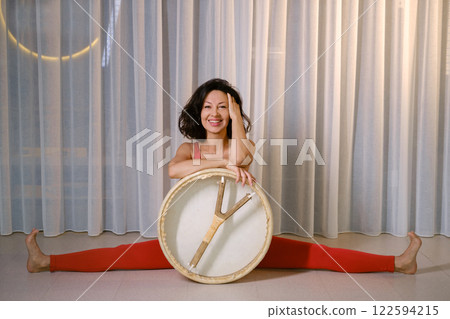 Woman in red tracksuit Sitting on a twine with a drum in a gym. Kalimeditation class Woman in red tracksuit Sitting on a twine with a drum in a gym. Kalimeditation class 122594215