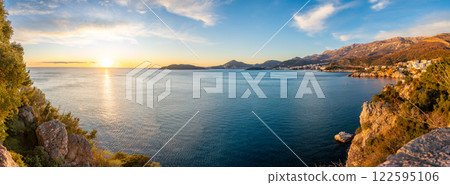 Coastal view of Becici and Budva from a rocky cliff, bathed in golden sunset light with the sea stretching toward the horizon. 122595106