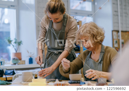 Two women work together with clay, developing skills and enjoying the artistic process. Elderly Caucasian woman learns pottery at a creative workshop with a young mentor 122596692