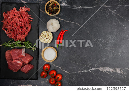 Top view of chunks of beef lying on a black cutting board, separated by sprigs of rosemary with red minced meat, vegetables and seasonings on a dark background with space for text. The concept of Top view of chunks of beef lying on a black cutting board, separated by sprigs of rosemary with red minced meat, vegetables and seasonings on a dark background with space for text. The concept of 122598527