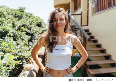 A cheerful young woman poses confidently outdoors by a wooden staircase, surrounded by lush greenery 122599097