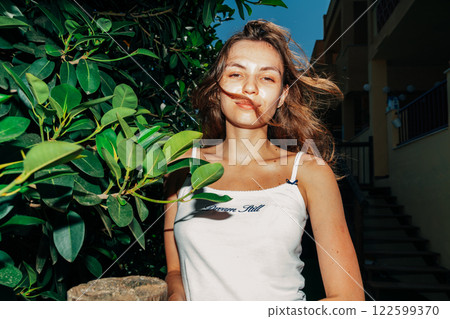 A young woman stands among lush green leaves, her hair flowing in the breeze, exuding confidence and A young woman stands among lush green leaves, her hair flowing in the breeze, exuding confidence and 122599370