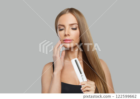 Posing young female model woman with long brown smooth hairstyle holding straight iron. 122599662