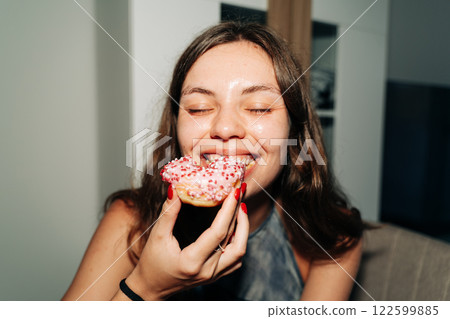 A cheerful young woman bites into a pink-frosted donut topped with sprinkles, enjoying a sweet treat A cheerful young woman bites into a pink-frosted donut topped with sprinkles, enjoying a sweet treat 122599885