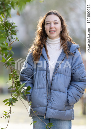 Young woman wearing blue puffer jacket is posing near green leaves Young woman wearing blue puffer jacket is posing near green leaves 122600011