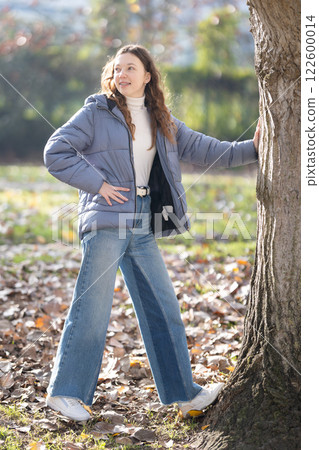 Young woman leaning against tree in autumn park enjoying sunny day 122600014