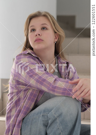 young woman sits thoughtfully on a staircase, her gaze directed into the distance. The image captures a quiet moment of reflection in a minimalist indoor setting. 122601551