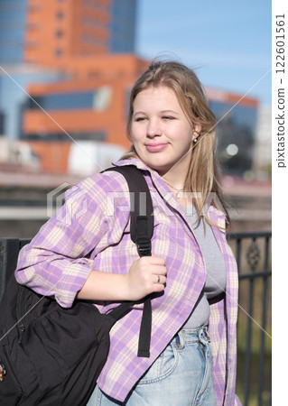 blonde girl, dressed casually, looks content as she stands outdoors with a bright cityscape behind her. This photo captures the essence of modern youth and freedom. 122601561