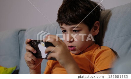 A boy is sitting on a sofa, visibly thrilled while playing on his smartphone. The image reflects a carefree moment of gaming fun and youthful enthusiasm. A boy is sitting on a sofa, visibly thrilled while playing on his smartphone. The image reflects a carefree moment of gaming fun and youthful enthusiasm. 122601583
