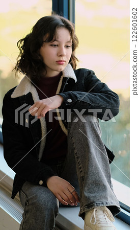 A contemplative girl sits by a window, her gaze directed outward. The photo highlights a reflective mood, symbolizing personal growth and the beauty of stillness. 122601602