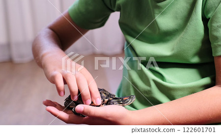 A close-up of a boy holding a turtle in his hand while gently petting its shell. The scene conveys a connection between a child and nature, showcasing care and curiosity. 122601701