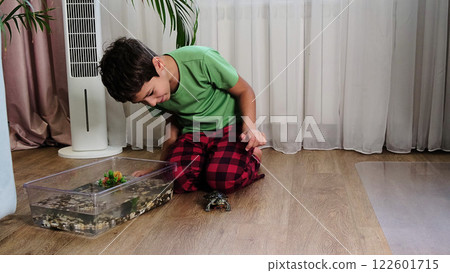 young boy kneels next to his turtle, which is on the floor near its habitat tank filled with water and aquatic decor. The image reflects the curiosity and care that children can have for their pets. 122601715