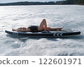 Woman in swimsuit basking in sun lying on sup board on frozen ice on river lake. 122601971
