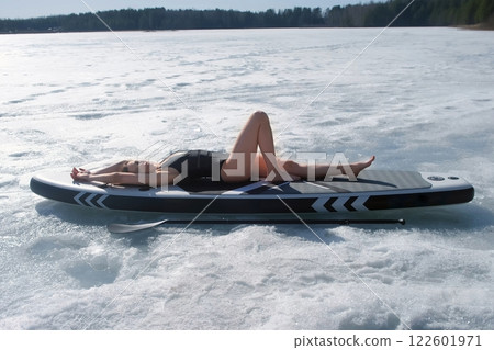 Woman in swimsuit basking in sun lying on sup board on frozen ice on river lake. 122601971