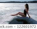 Woman in swimsuit basking in sun sitting on snow near ice hole on river. 122601972