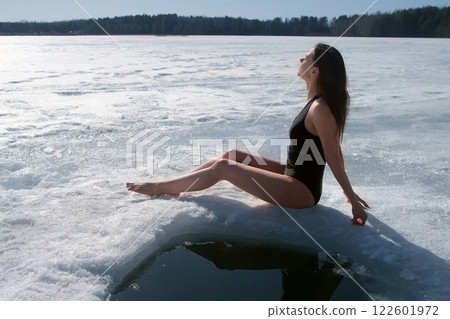 Woman in swimsuit basking in sun sitting on snow near ice hole on river. Woman in swimsuit basking in sun sitting on snow near ice hole on river. 122601972