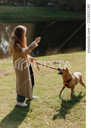young woman walks with her dog in the park. A lady in a beige raincoat is playing with a pit bull young woman walks with her dog in the park. A lady in a beige raincoat is playing with a pit bull 122602140