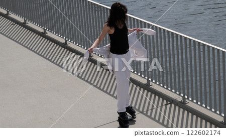 A stylish woman pauses near the water, holding her jacket while standing on rebound shoes. The combination of fitness and fashion highlights modern trends in active and mindful living. 122602174