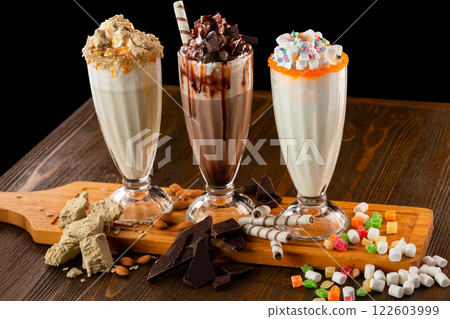 Three sweet milkshakes with chocolate, marshmallow, halva and whipped cream at a wooden board on a table isolated at black background. Three sweet milkshakes with chocolate, marshmallow, halva and whipped cream at a wooden board on a table isolated at black background. 122603999
