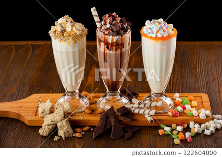 Three sweet milkshakes with chocolate, marshmallow, halva and whipped cream at a wooden board on a table isolated at black background. 122604000